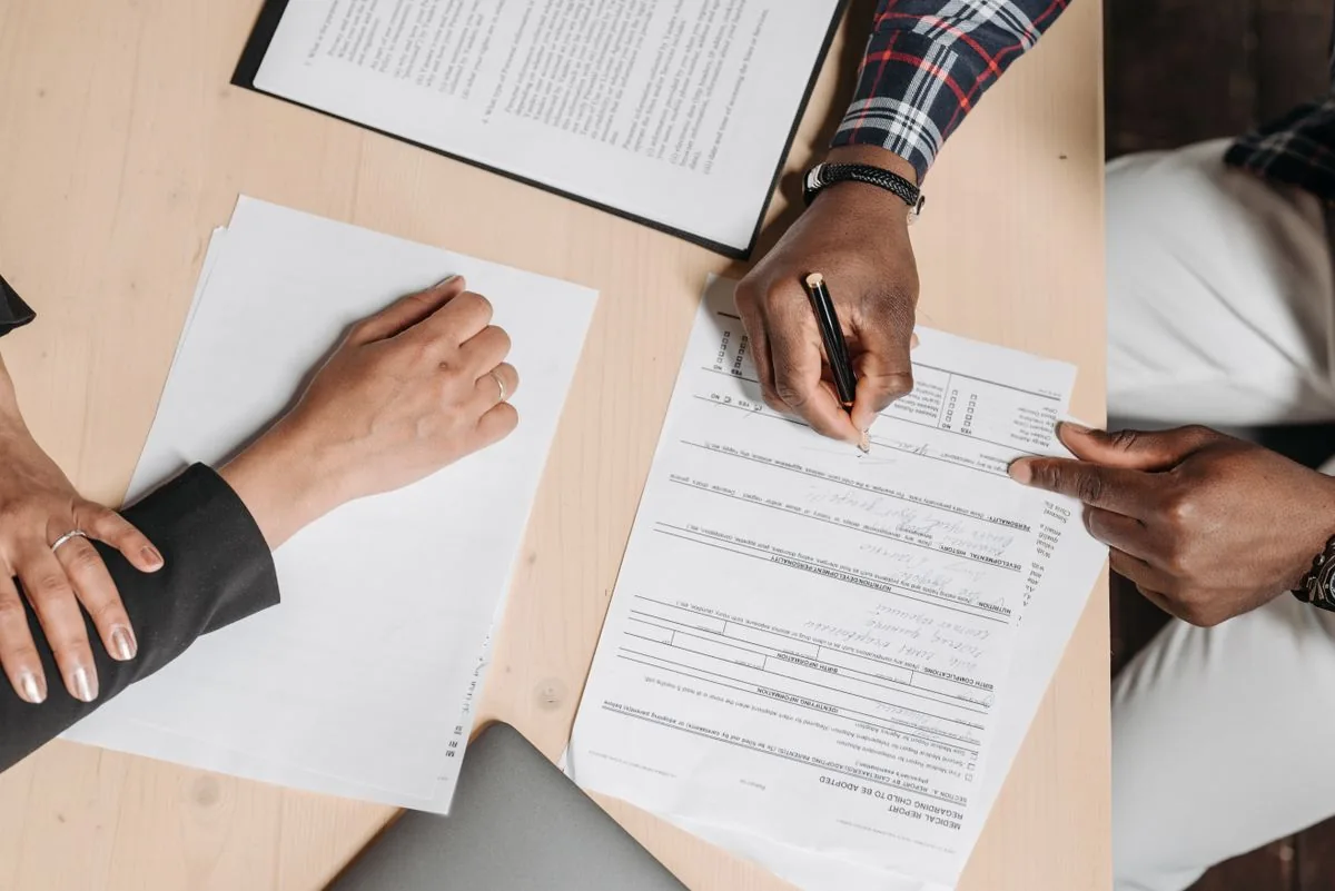 Two people review paperwork and sign documents at a desk.